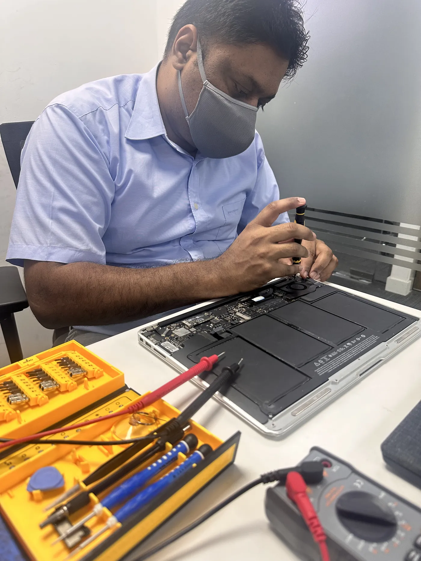 TechFix technician testing a MacBook logic board with a multimeter